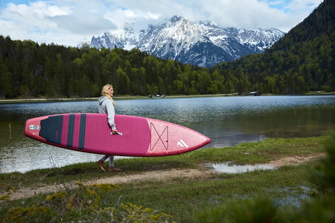 Duotone Diamond Air Touring inflatable SUP board 11'6" in raspberry pink carried at an alpine lake shore