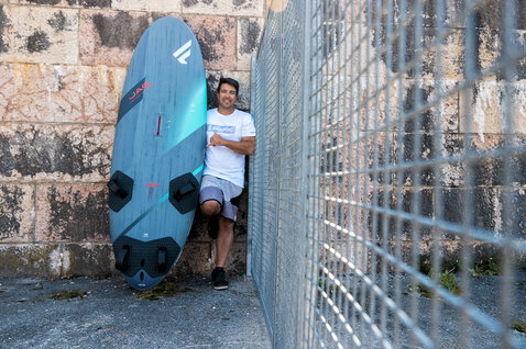 Fanatic Jag LTD windsurf board in slate teal held by rider against a stone wall